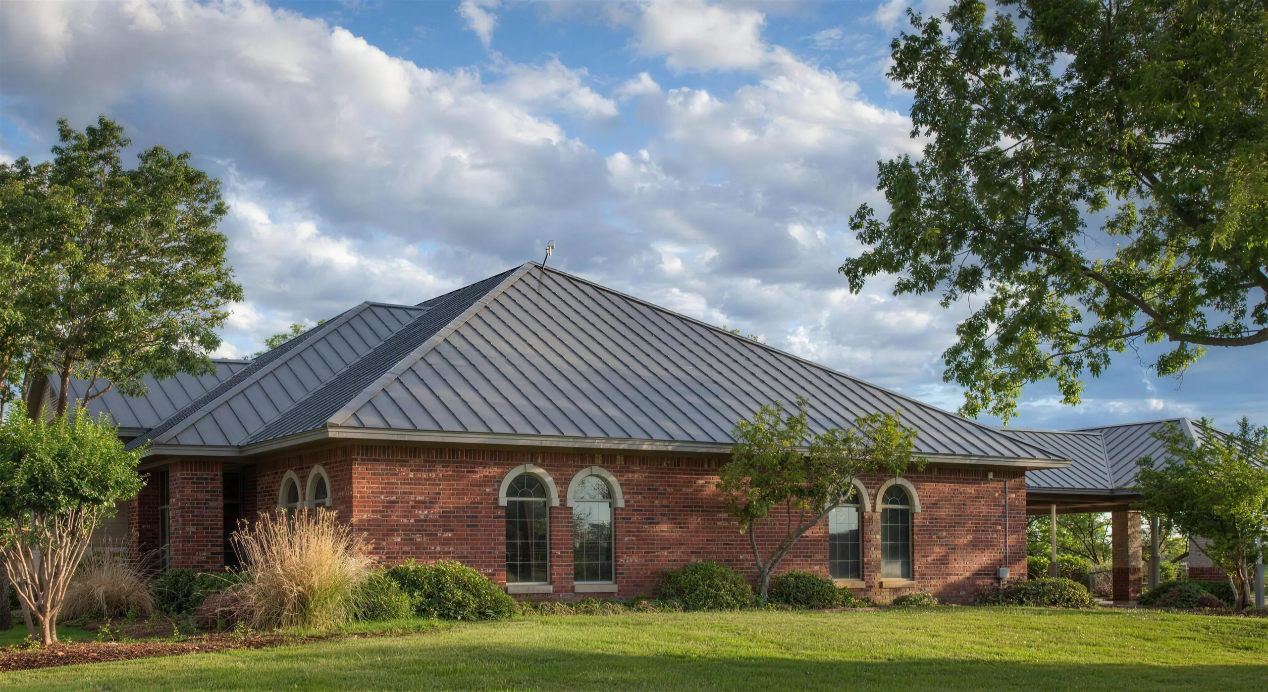 Red-brick house under a dark standing-seam metal roof, big white clouds over the ridge and late sun on the lawn.