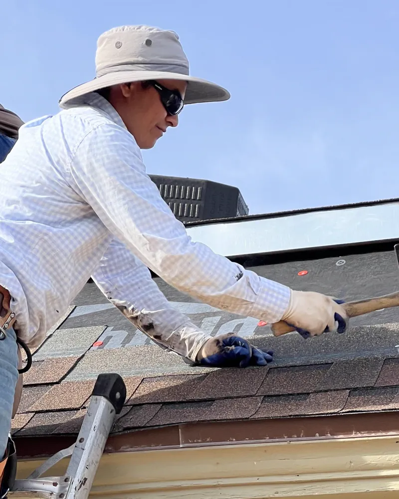 Roofer in a wide-brim sun hat nailing new shingles at the eave.